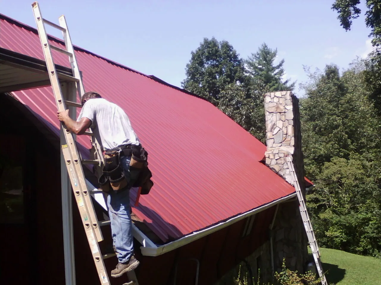 Roofer on a ladder beside a red metal roof and stone chimney — Soffit & Fascia Repair by Roofing Brothers in West Lafayette